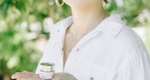 Young woman in white shirt holding a small container of CBD cream outdoors in sunlight.