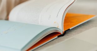 A person flips through a book beside a laptop on a desk, suggesting study or work.