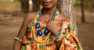 Portrait of a girl in Kente cloth and jewelry, set against a natural outdoor background.