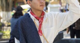 Young man in traditional Mexican clothing holding a sombrero in a sunny outdoor plaza.