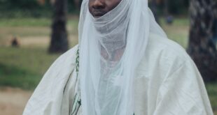 Man in traditional African clothing and headdress holding a sword outdoors with palm trees.
