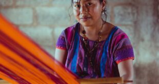 Indigenous Mexican woman weaving colorful fabric on a traditional loom inside a rustic setting.