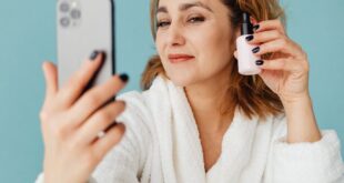 Woman in a white robe taking a selfie with skincare products on table.