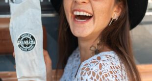Cheerful woman showcasing shawarma while smiling, wearing a white top and black hat.