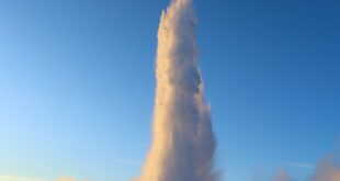 Dramatic geyser eruption in Iceland with bright blue sky and snowy foreground.