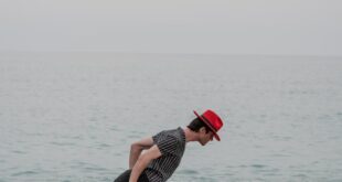 A man in a striped shirt and red hat leans in a unique pose on a sandy beach.