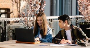 Two students collaborate on a laptop at an outdoor campus setting with cherry blossoms.