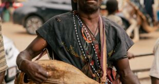 An African musician in traditional attire playing an instrument on a lively street.